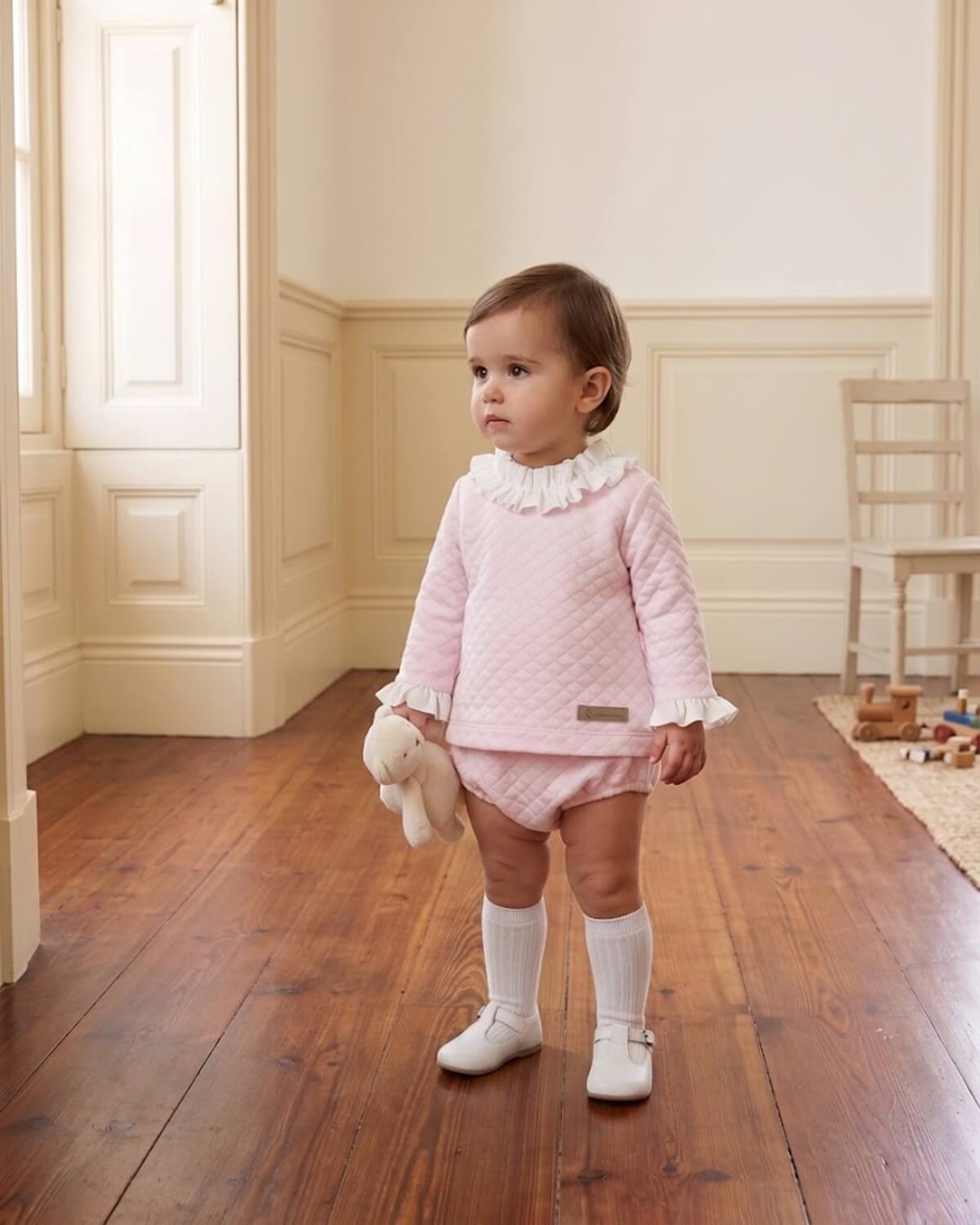 Baby girl standing wearing Calamaro traditional Spanish pink quilted two-piece set with ruffle collar, bloomers and white knee socks.