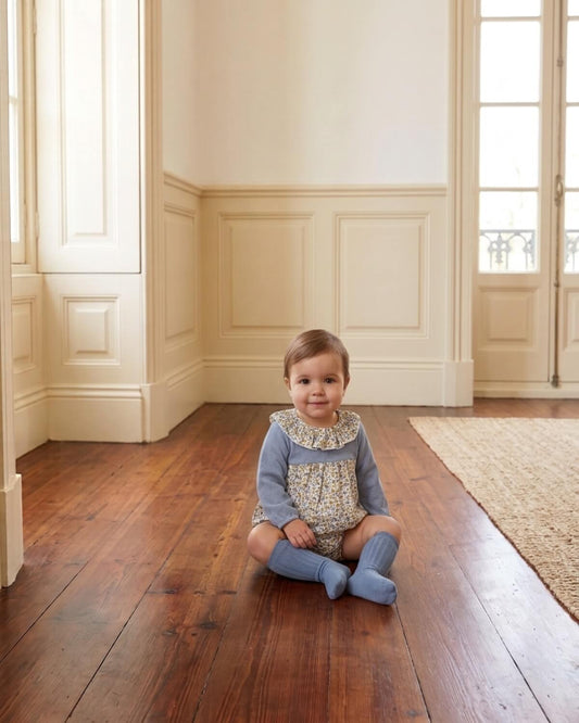 Baby girl sitting wearing traditional Spanish blue floral romper with frilled collar and matching knee socks.