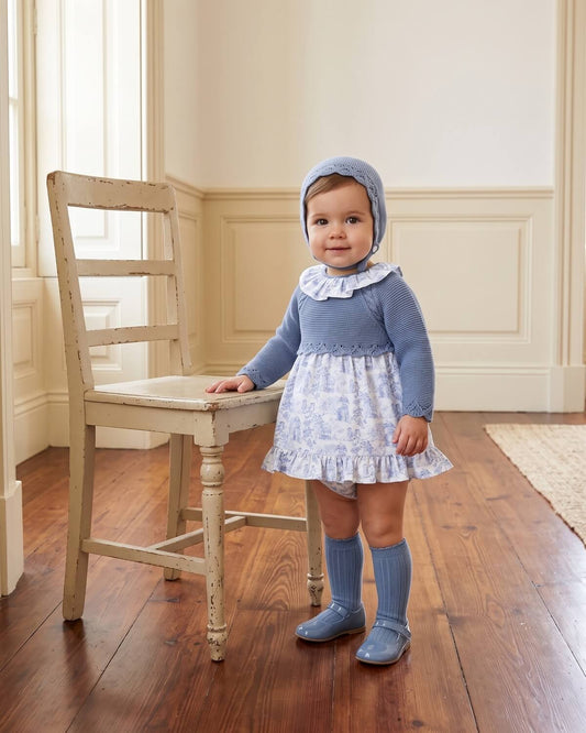 Baby girl wearing traditional Spanish dusty blue knitted dress with toile de Jouy skirt, matching bonnet and knee socks.
