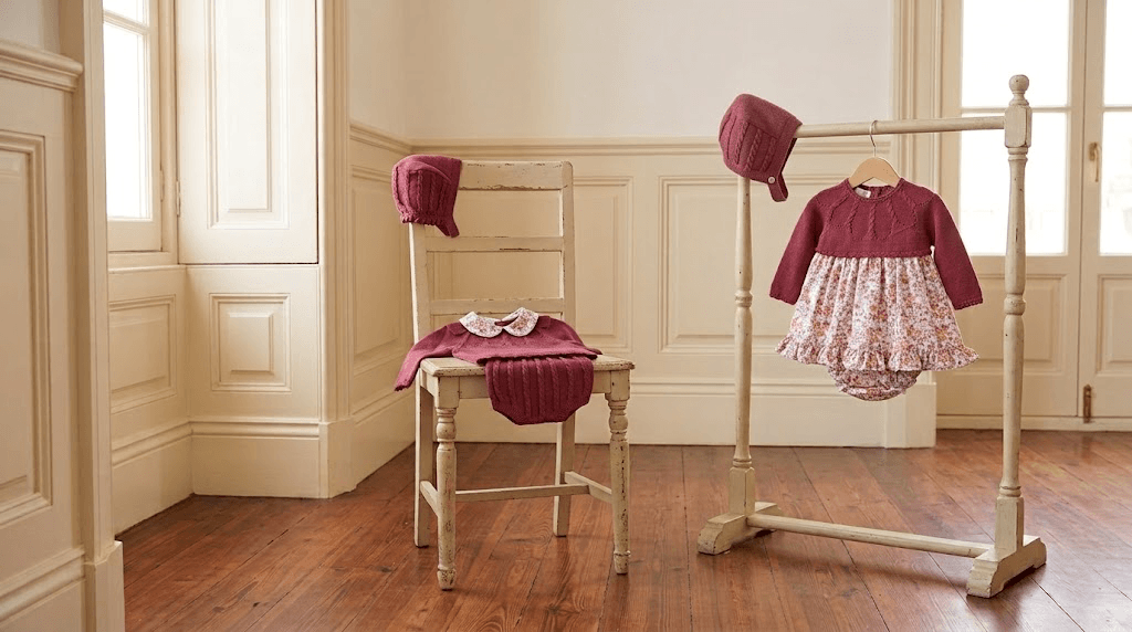 Matching Dulce de Fresa traditional Spanish baby collection in burgundy knit and floral print displayed on a vintage chair and wooden clothing rack.